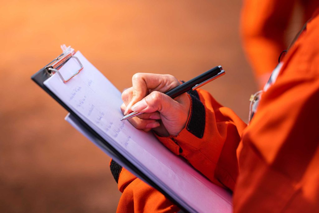 Mulher em jaleco laranja tomando notas sobre sinalização de NR-12 em prancheta.